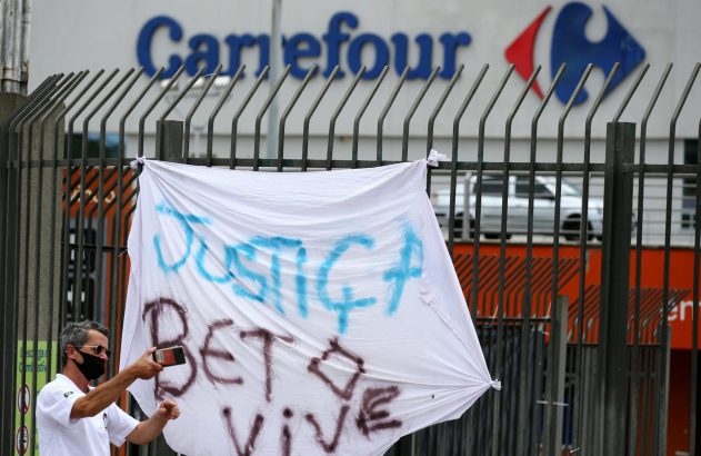 Faixa de protesto "JUSTIÇA BETO VIVE" em frente a um Carrefour, com homem tirando foto.