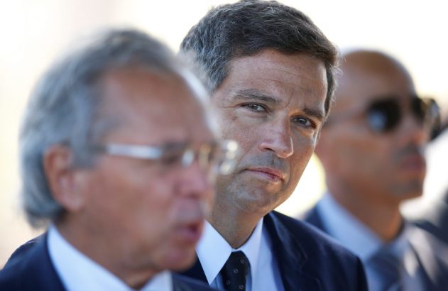 Um homem em terno, em foco, olha à frente, com outro homem desfocado em primeiro plano e um terceiro ao fundo.