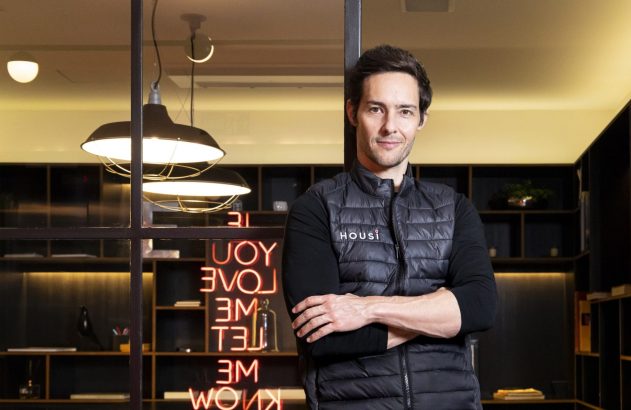 Homem sorridente de colete Housi, braços cruzados, em escritório moderno com luminárias industriais e letreiro neon.