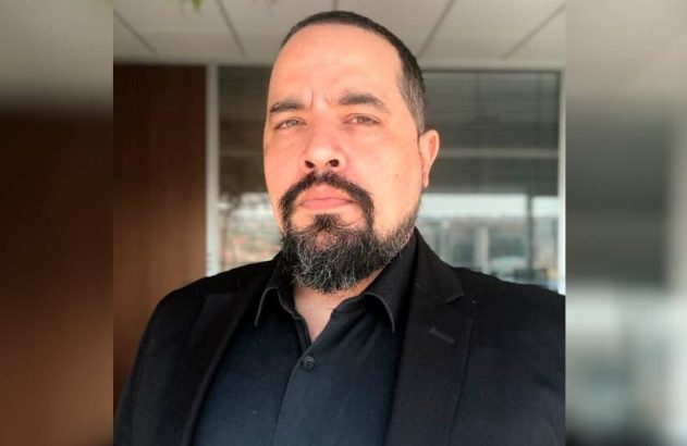 Homem barbudo de blazer preto olhando para a câmera. Fundo interno desfocado.