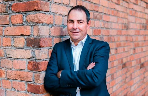 Homem de terno, braços cruzados, sorrindo contra uma parede de tijolos.