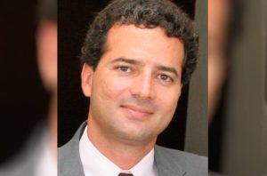 Retrato de um homem sorrindo, de terno e cabelo escuro encaracolado.