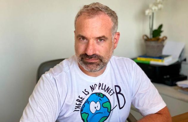 Homem sentado à mesa, vestindo camiseta branca com a frase 'THERE IS NO PLANET B', olhando para a câmera.