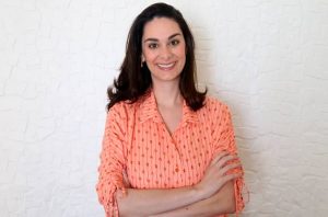 Mulher sorrindo, braços cruzados, vestindo camisa pêssego estampada. Fundo branco texturizado.