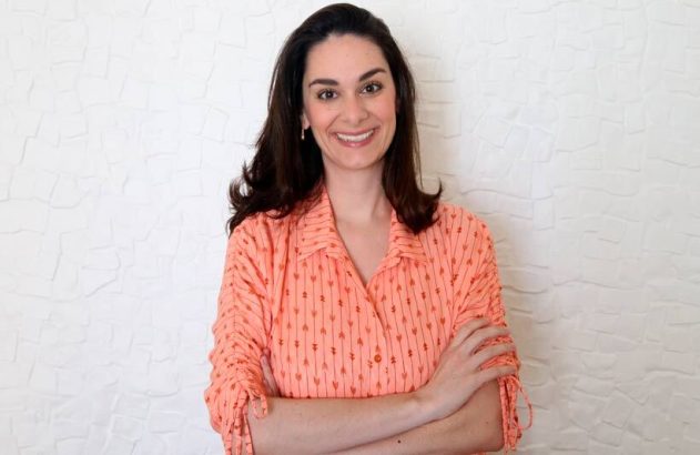 Mulher sorrindo, braços cruzados, vestindo camisa pêssego estampada. Fundo branco texturizado.