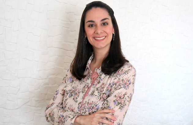 Mulher com cabelo escuro e camisa floral sorri, braços cruzados, em frente à parede branca texturizada.