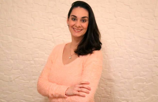 Mulher sorrindo, de suéter rosa claro felpudo e braços cruzados, em frente a uma parede texturizada.