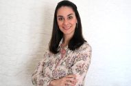 Mulher sorridente com cabelos escuros e camisa floral, braços cruzados, em pé contra parede branca texturizada.