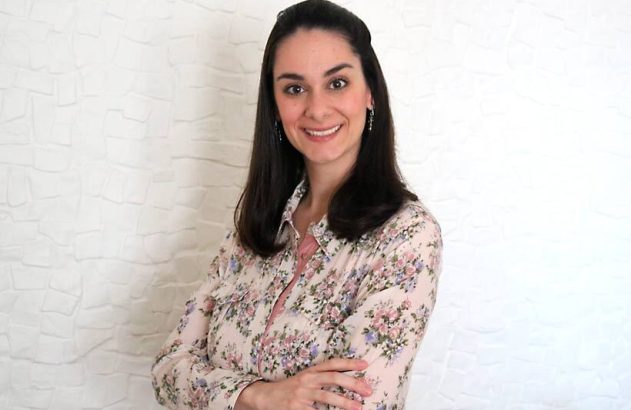 Mulher sorridente com cabelos escuros e camisa floral, braços cruzados, em pé contra parede branca texturizada.