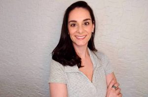 Mulher de cabelo escuro sorrindo e olhando para a câmera, com braços cruzados e blusa cinza, em fundo de parede texturizada.