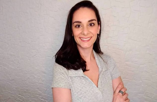 Mulher de cabelo escuro sorrindo e olhando para a câmera, com braços cruzados e blusa cinza, em fundo de parede texturizada.