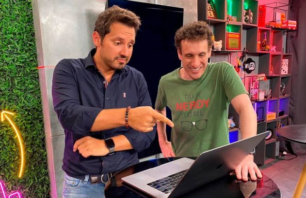 Dois homens olham para um laptop; um aponta e o outro sorri em um estúdio decorado.