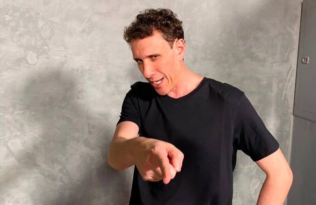 Homem de camiseta preta, com expressão energética, apontando para frente e para baixo.