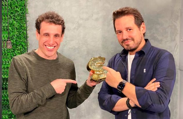 Dois homens sorridentes, um segurando e ambos apontando para um prêmio dourado estilizado com a letra S.