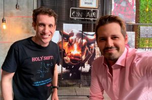 Dois homens sorrindo posam juntos, um de camiseta escura e outro de camisa clara. Há uma imagem de fogo no fundo.