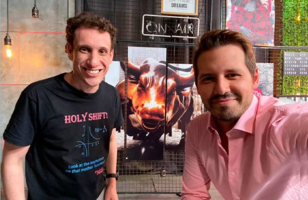 Dois homens sorrindo posam juntos, um de camiseta escura e outro de camisa clara. Há uma imagem de fogo no fundo.