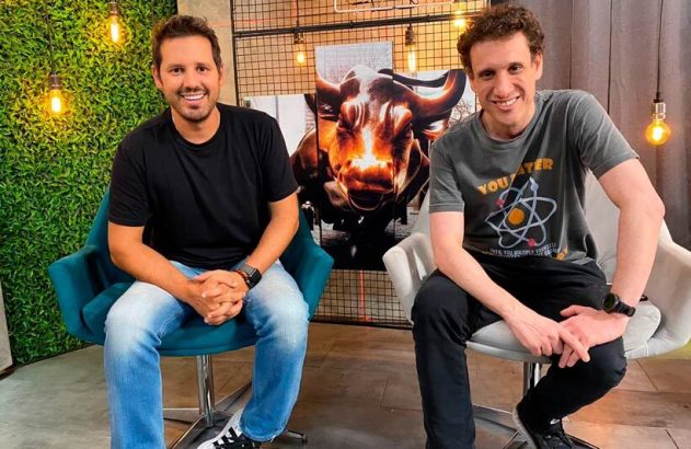 Dois homens sorrindo sentados em um estúdio moderno com arte de touro ao fundo.