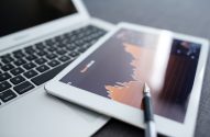 Gráfico de dados financeiros em tablet, com caneta e laptop ao fundo.