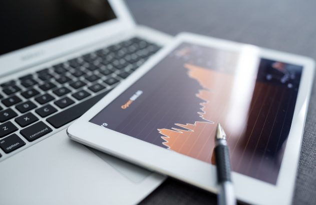 Gráfico de dados financeiros em tablet, com caneta e laptop ao fundo.