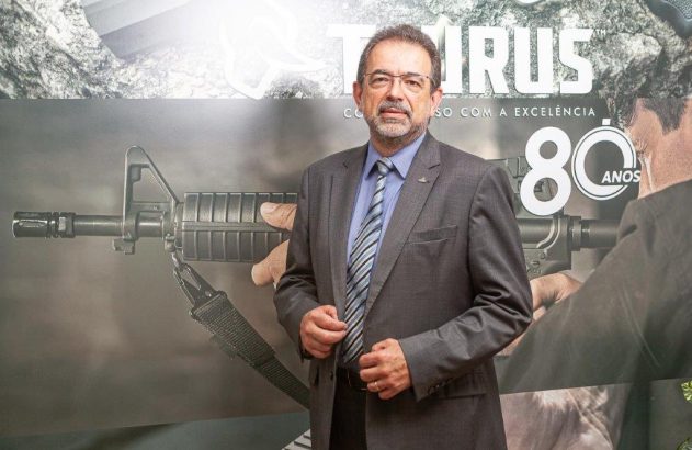 Homem de terno olhando para a câmera em frente a um pôster de rifle e texto.