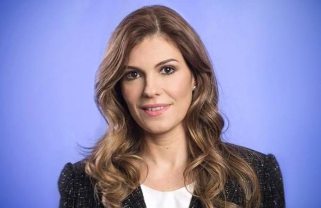 Mulher sorrindo com cabelo castanho e blazer preto em frente a um fundo azul.