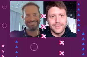 Videochamada: dois homens. Um sorri, outro neutro. Fundo roxo com formas geométricas.