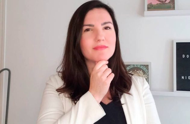 Mulher de cabelos escuros e blazer claro, com a mão no queixo e expressão pensativa. Fundo claro e desfocado.