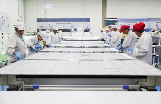 Trabalhadores montando painéis solares em linha de produção de fábrica.