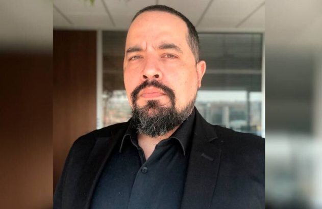 Homem barbudo de blazer preto olhando para a câmera. Fundo interno desfocado.