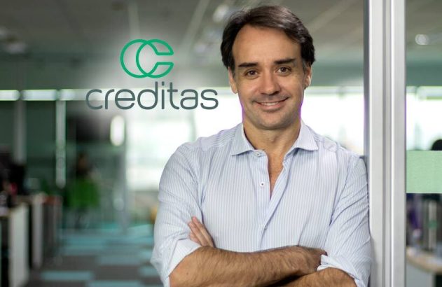 Homem sorrindo com braços cruzados em escritório, com logo Creditas no vidro.