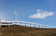 Grande cruz branca em colina seca com cerca, sob céu azul com nuvem.