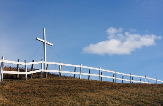 Grande cruz branca em colina seca com cerca, sob céu azul com nuvem.