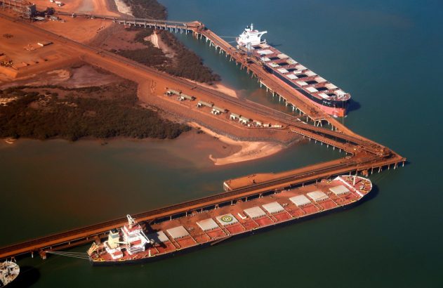 Vista aérea de terminal de minério com dois navios de carga a granel atracados, e esteiras transportadoras no cais.