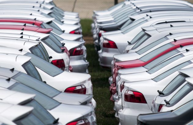 Duas longas filas paralelas de carros brancos, prateados e vermelhos, vistos de traseira em um pátio.