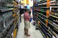 Mulher idosa olhando bebidas em um corredor de supermercado, segurando uma pequena cesta.
