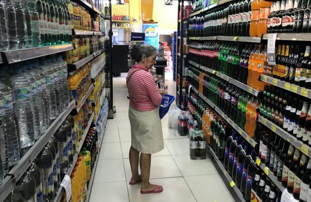 Mulher idosa olhando bebidas em um corredor de supermercado, segurando uma pequena cesta.