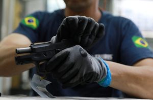 Pessoa com luvas manuseia pistola Taurus. Na manga, patch da bandeira do Brasil.