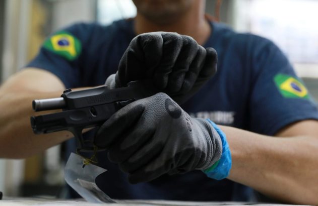Pessoa com luvas manuseia pistola Taurus. Na manga, patch da bandeira do Brasil.