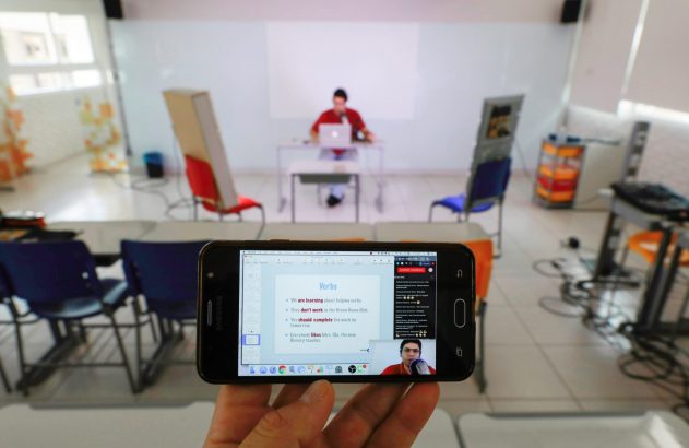 Mão segura smartphone com aula online de inglês: slides de verbos e professor. Sala de aula vazia no fundo.