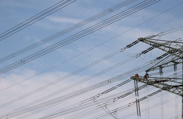 Dois trabalhadores em uma torre de alta tensão, realizando manutenção em linhas elétricas sob um céu azul.