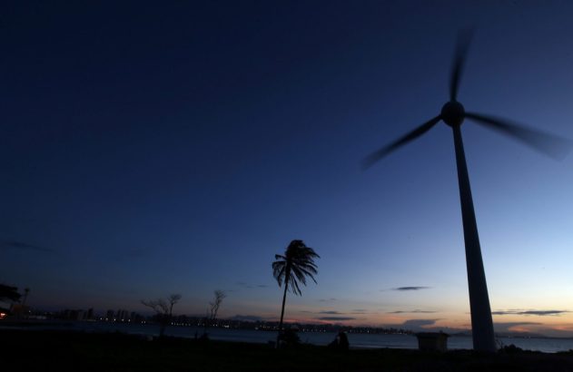 Anoitecer: céu azul escuro, gerador eólico de pás borradas à direita, palmeira e cidade litorânea iluminada ao fundo.