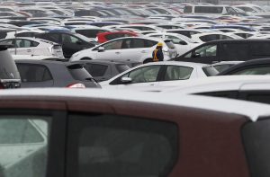 Pátio de estacionamento lotado de carros novos. Um trabalhador com capacete e colete caminha entre os veículos.