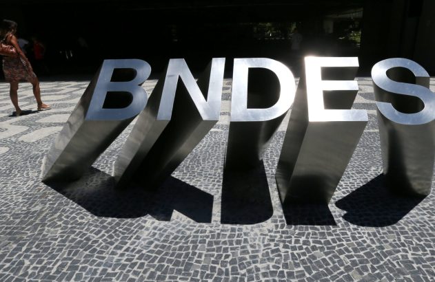 Letras metálicas "BNDES" projetam sombras longas em calçamento de pedras. Uma mulher caminha ao fundo.
