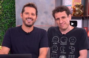 Dois homens em um estúdio. O da esquerda sorri. O da direita tem expressão neutra e veste camiseta com desenhos.