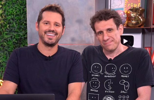 Dois homens em um estúdio. O da esquerda sorri. O da direita tem expressão neutra e veste camiseta com desenhos.