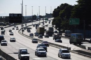 Rodovia movimentada com vários veículos e caminhões. Sinalização para Barra, Centro, Copacabana visível à direita.