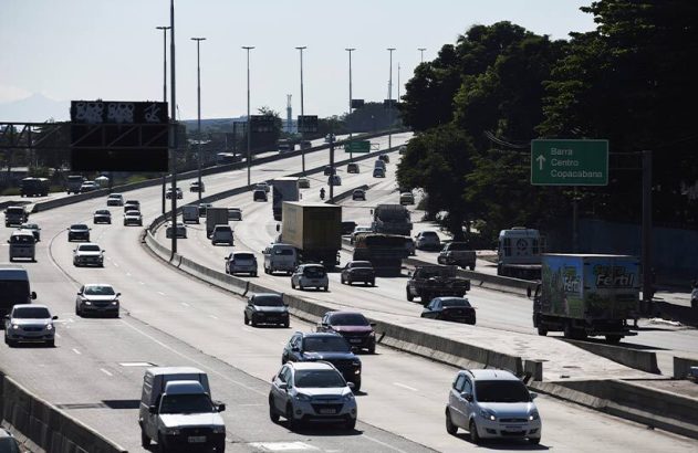 Rodovia movimentada com vários veículos e caminhões. Sinalização para Barra, Centro, Copacabana visível à direita.