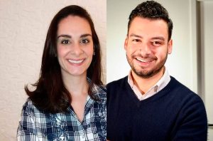 Um homem e uma mulher sorrindo para a câmera. Ela de camisa xadrez; ele de suéter azul e barba.