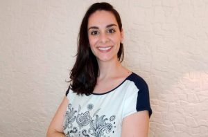 Mulher sorridente com cabelos escuros e camiseta branca estampada, em frente a uma parede clara e texturizada.