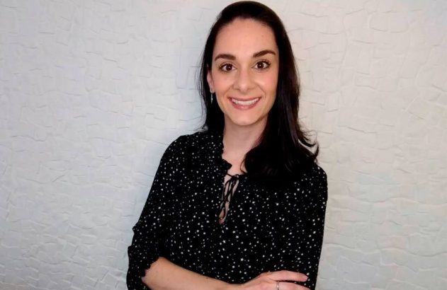 Mulher sorridente de cabelo escuro, blusa escura de bolinhas e braços cruzados em fundo claro.